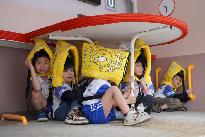 《圖說》「健康安全，幸福成長－115年新北市兒童安全宣導活動」，小朋友在地震體驗車內戴保護頭套躲在桌下。〈社會局提供〉